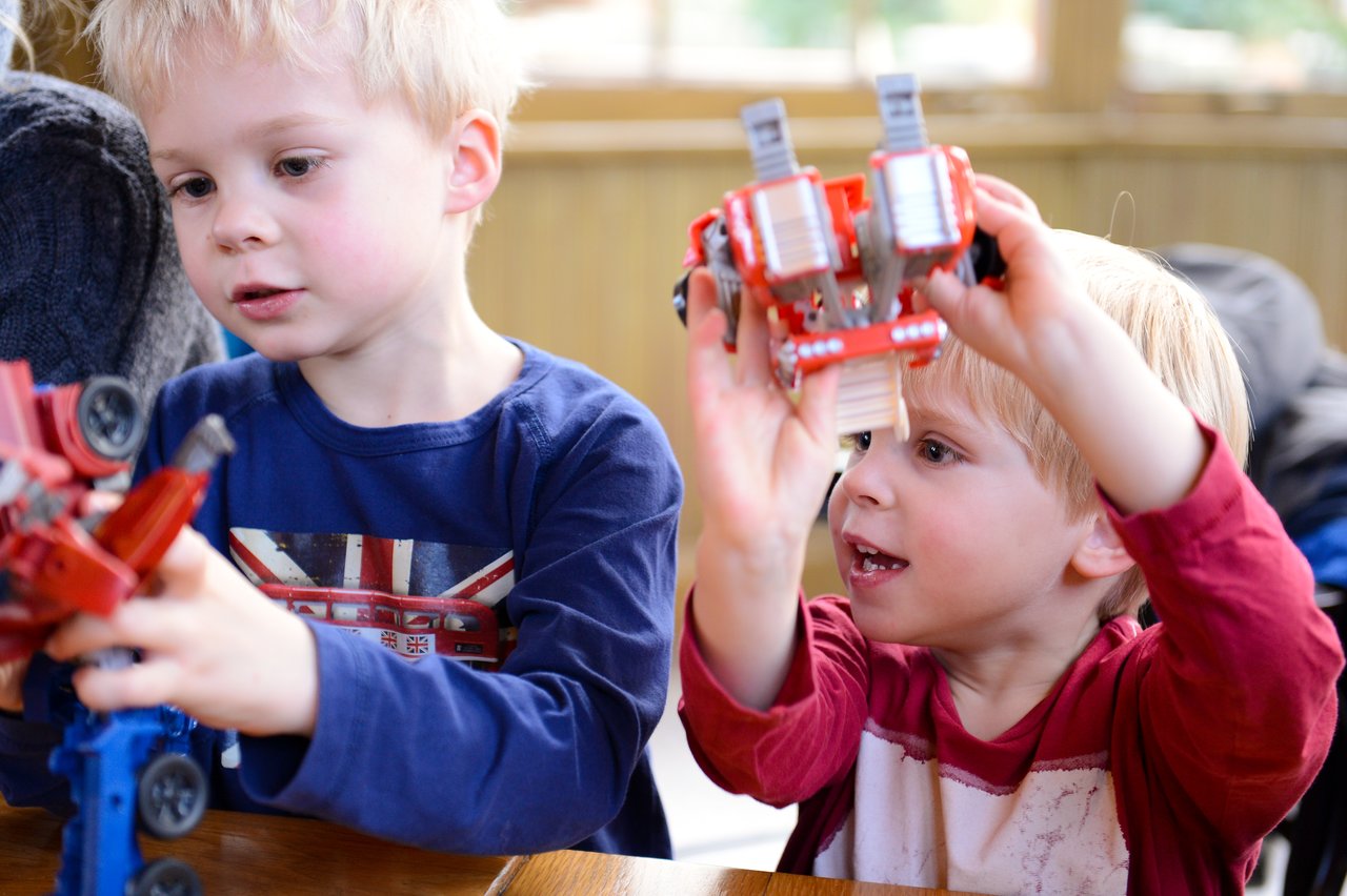 Two young children play with red Transformer toys, one holding it up while the other examines it closely.