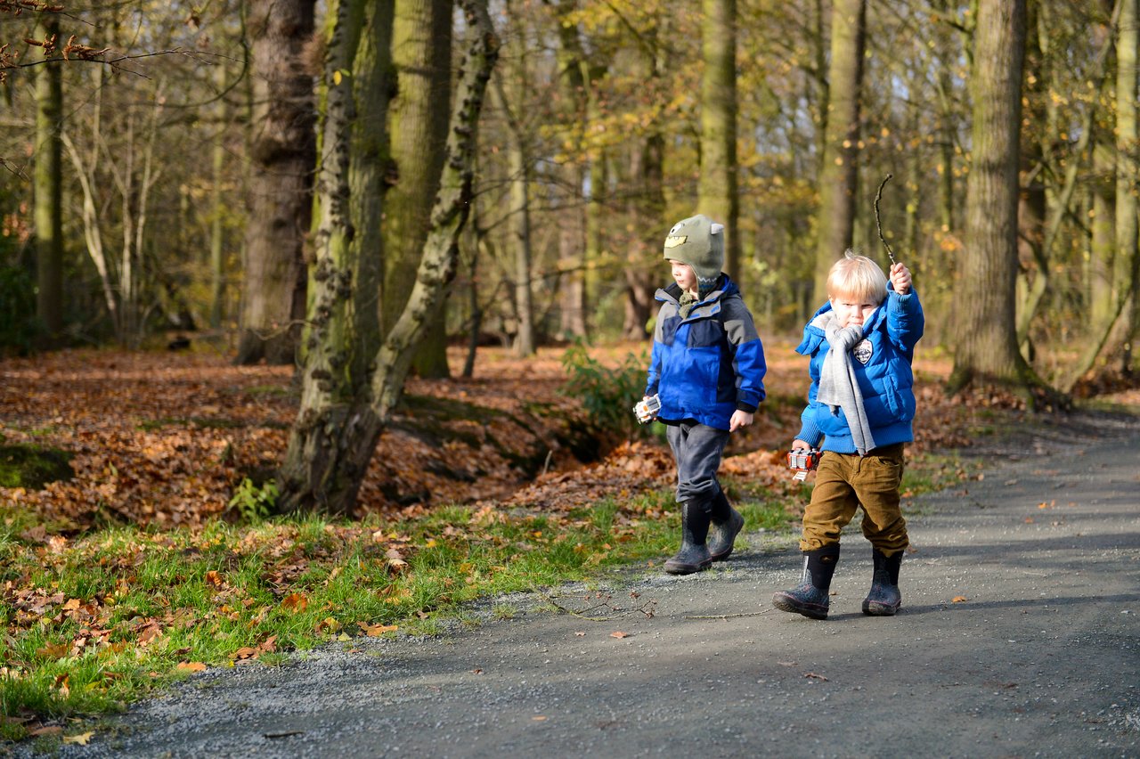 Two young children in warm clothes walk along a park path, one holding up a stick.