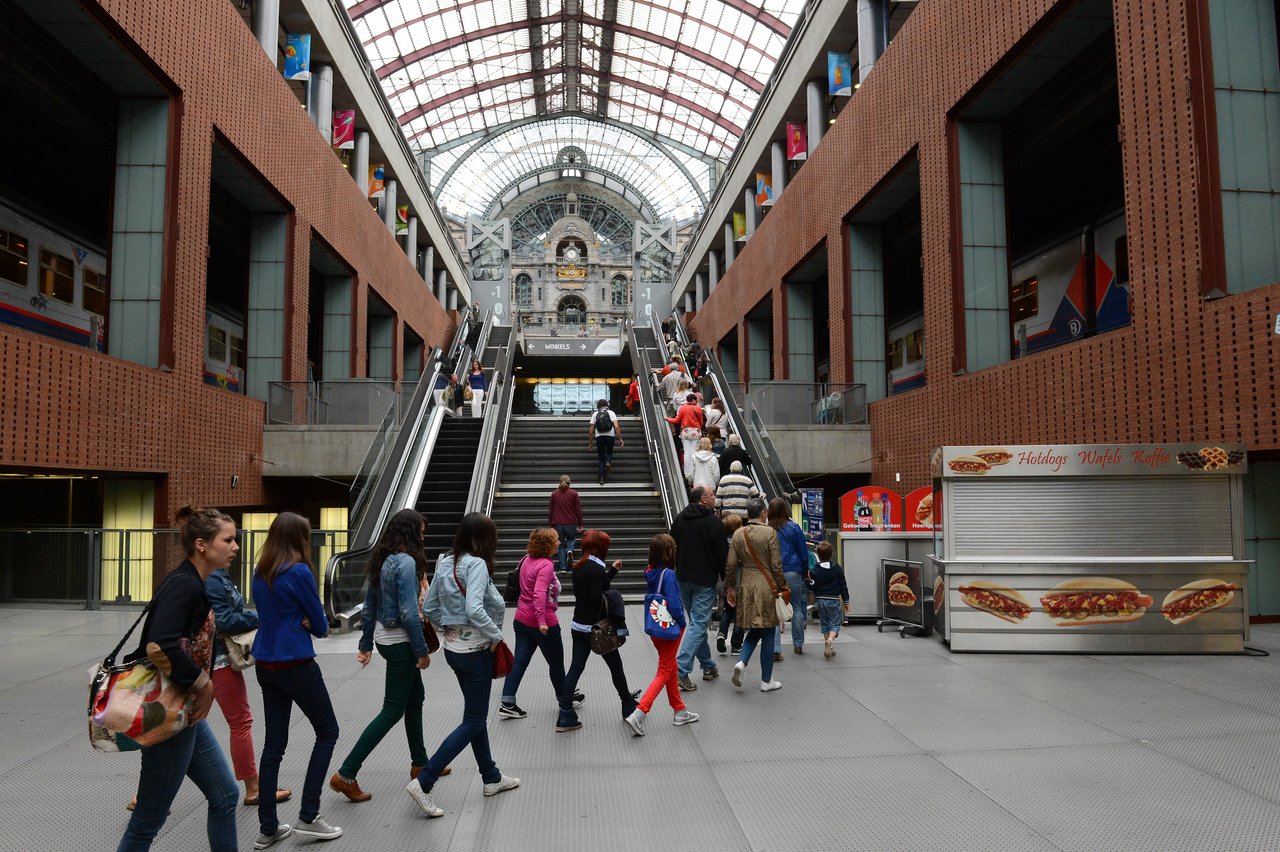 People walk through Antwerp Central Station, some using escalators and stairs, with trains visible on the upper level.