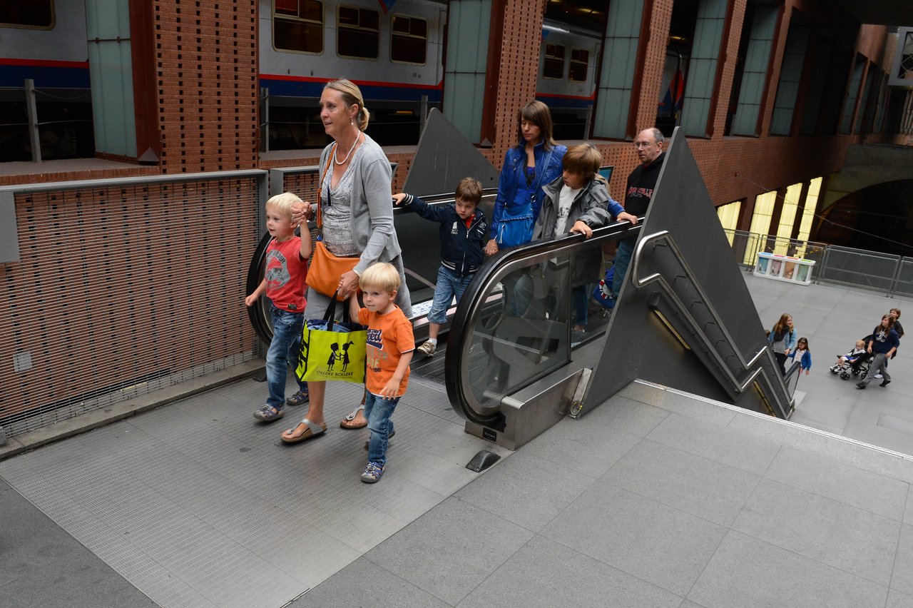 People, including children, walk near an escalator inside Antwerp Central Station, with a train visible in the background.