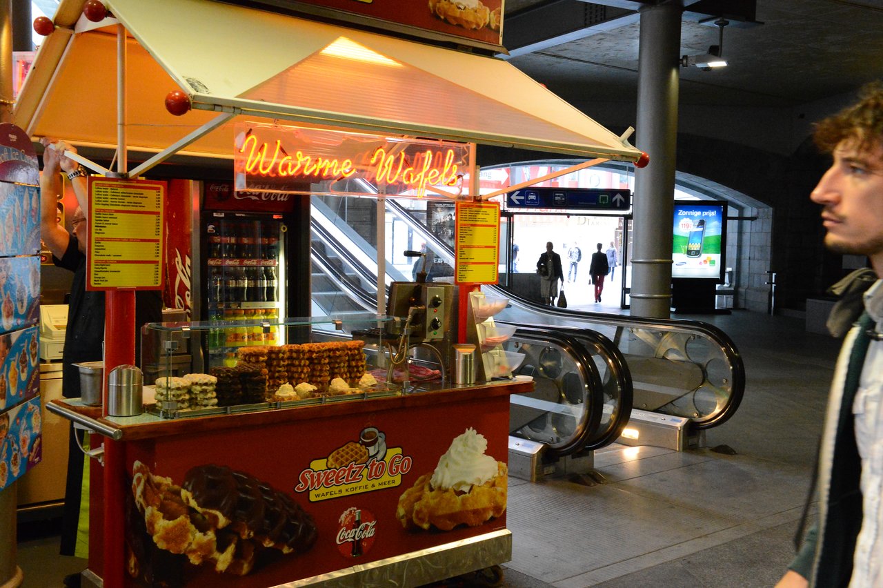A waffle stand at Antwerp Central Station with waffles on display and a neon sign reading "Warme Wafels.