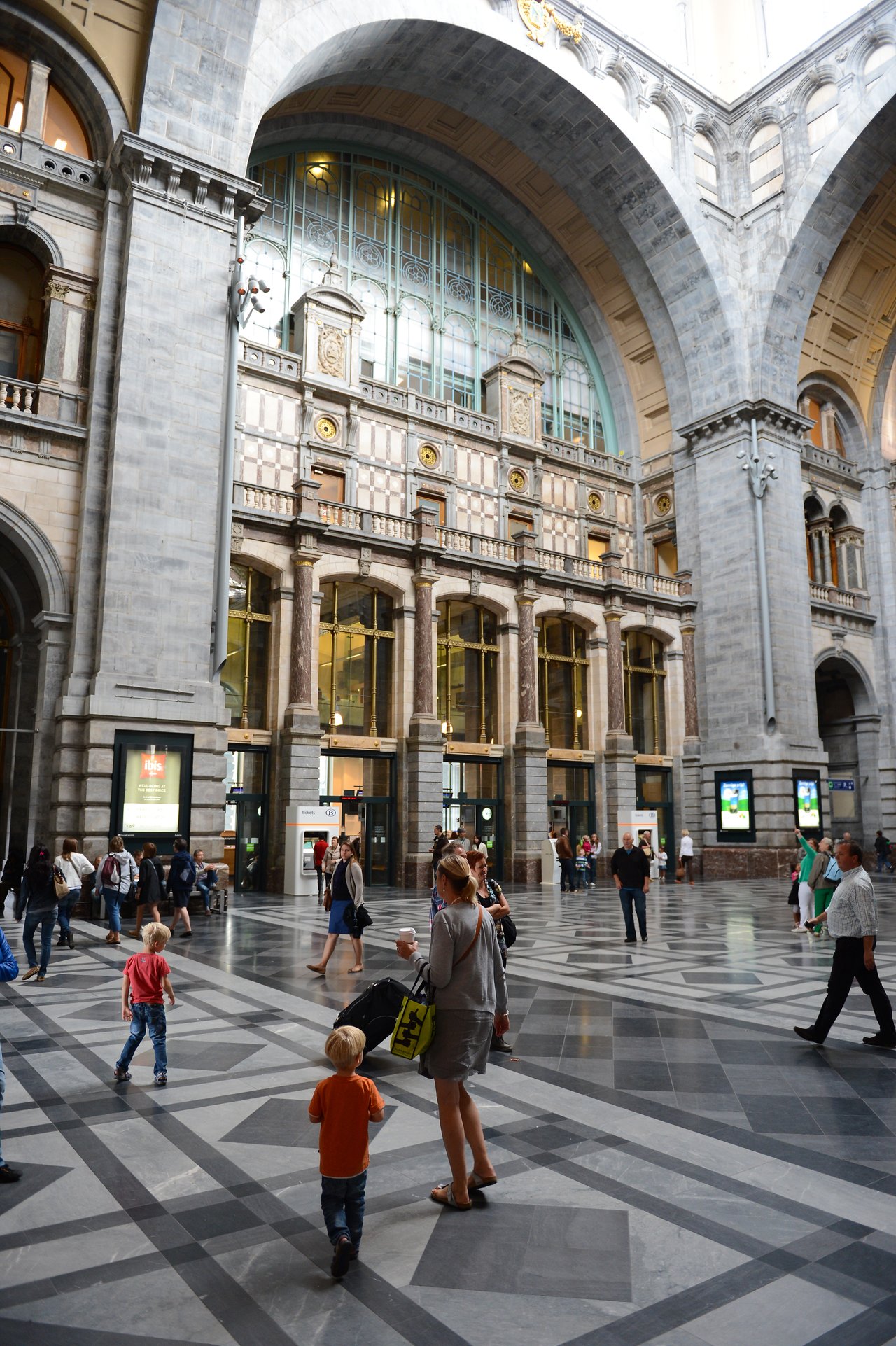 People walk through the spacious hall of Antwerp Central Station, with high arches and decorative windows in the background.