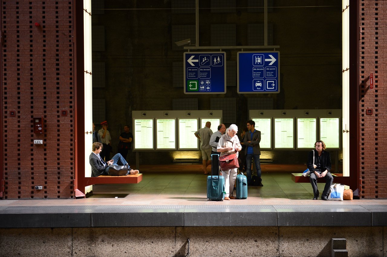 Passengers wait at Antwerp Central Station, some sitting and others standing with luggage, checking schedules and signs.