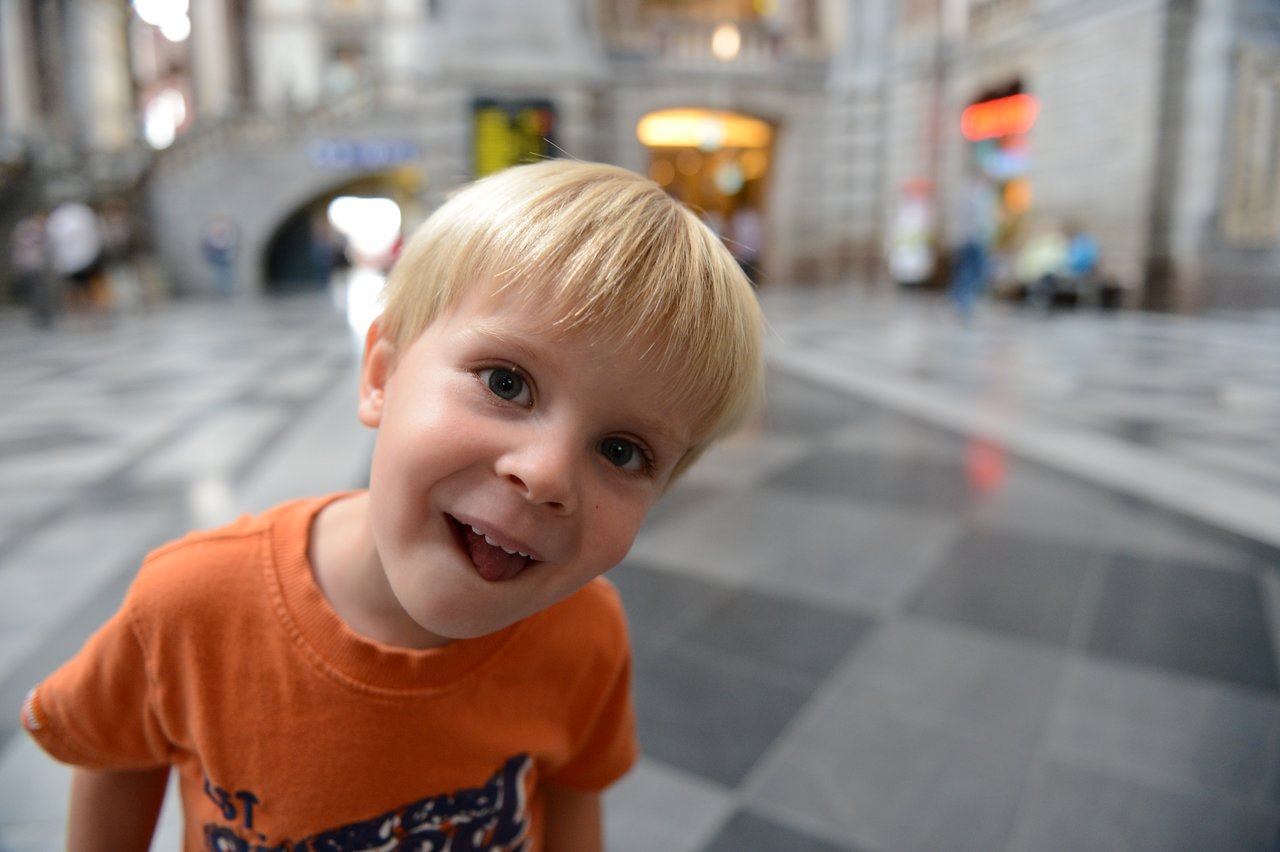 A young child in an orange shirt leans toward the camera, smiling playfully inside Antwerp Central Station.