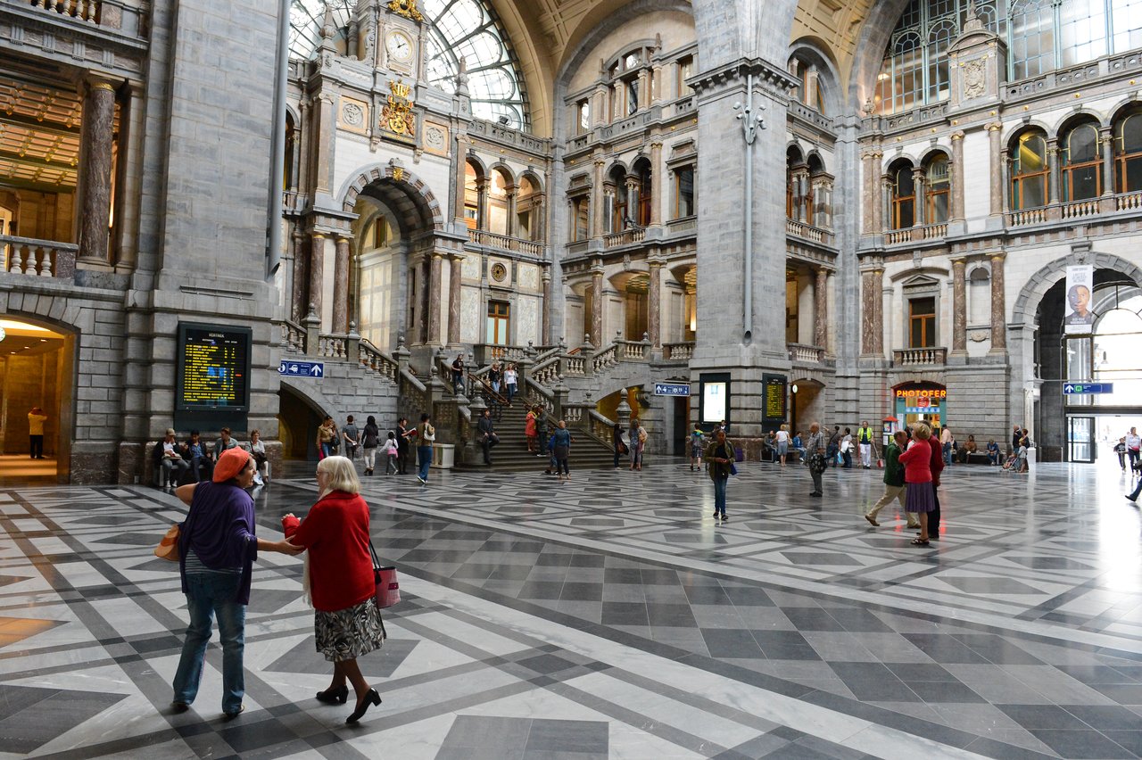 People walk and talk inside Antwerp Central Station, a grand historic building with high ceilings, arches, and staircases.