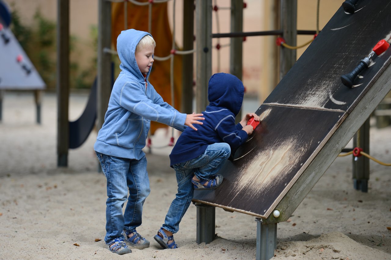 A child in a blue hoodie climbs a playground ramp while another child helps by pushing from behind.