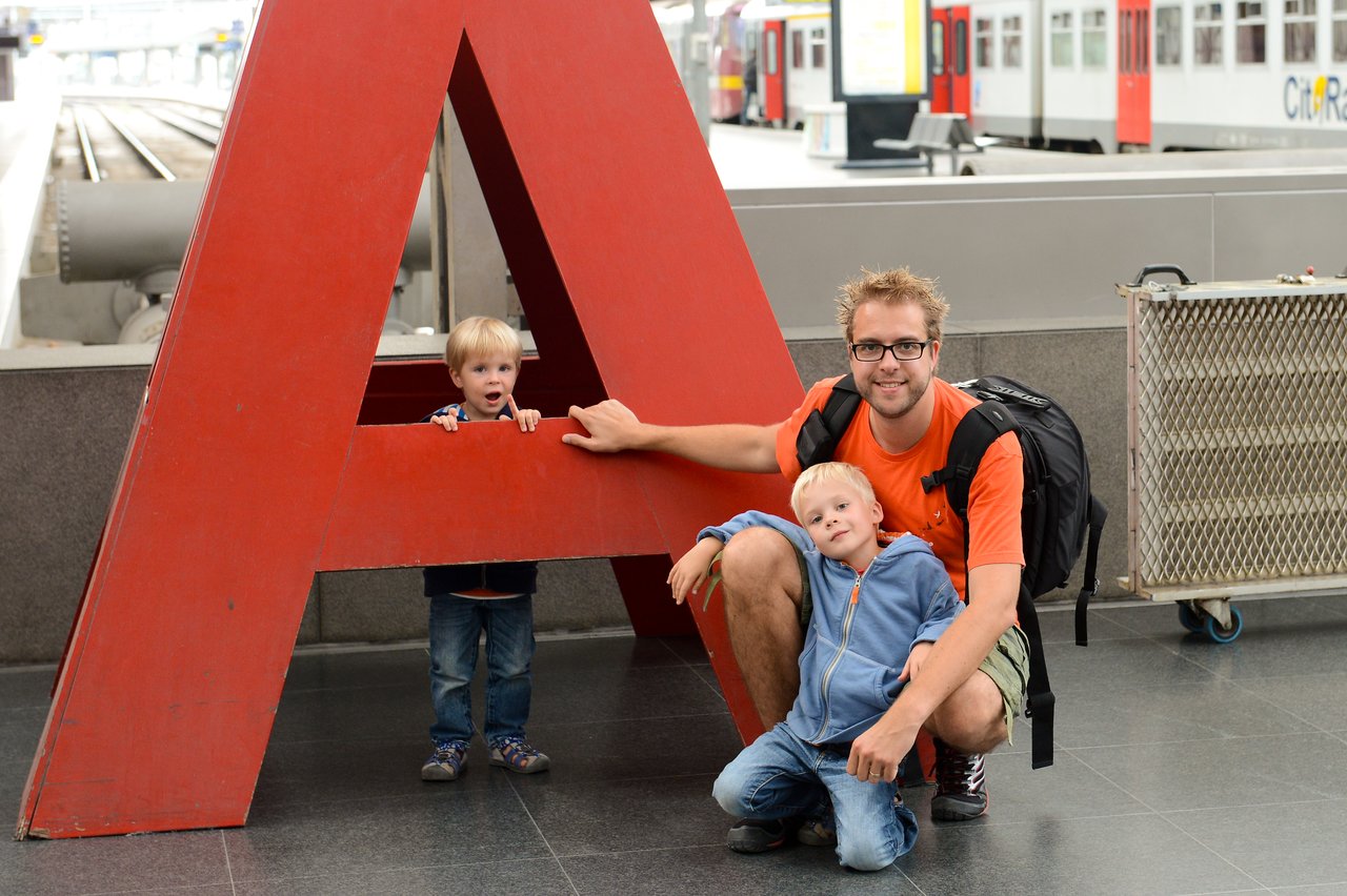 A man with a backpack and two children pose near a large red letter "A" at a train station.