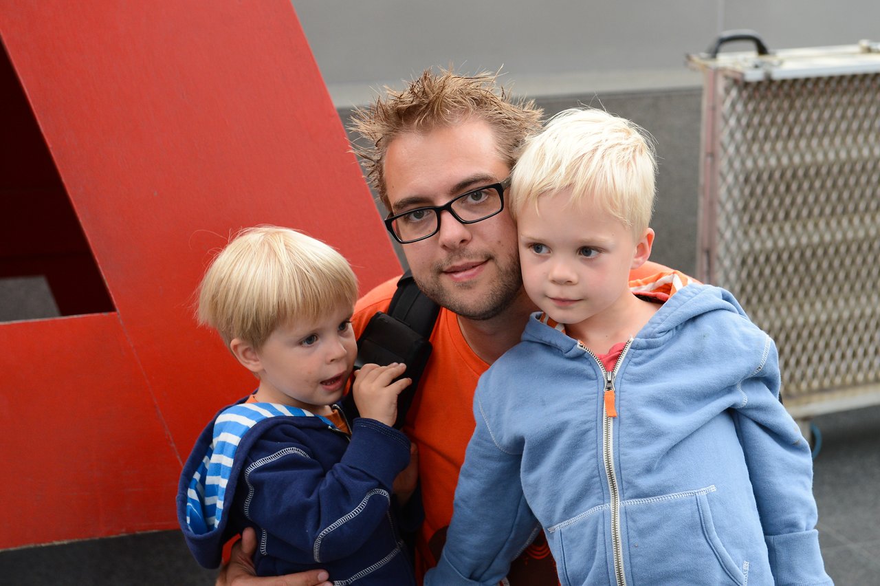 A man in glasses and an orange shirt poses with two young children wearing blue hoodies.