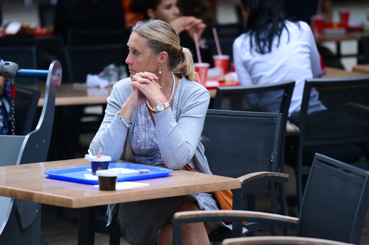 A woman sits at an outdoor table, hands clasped, with a tray holding coffee and dessert in front of her.