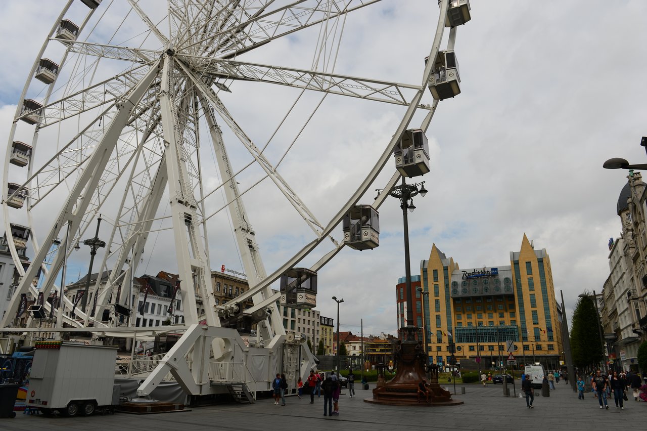 A large Ferris wheel with enclosed cabins stands in a public square, with people walking nearby.