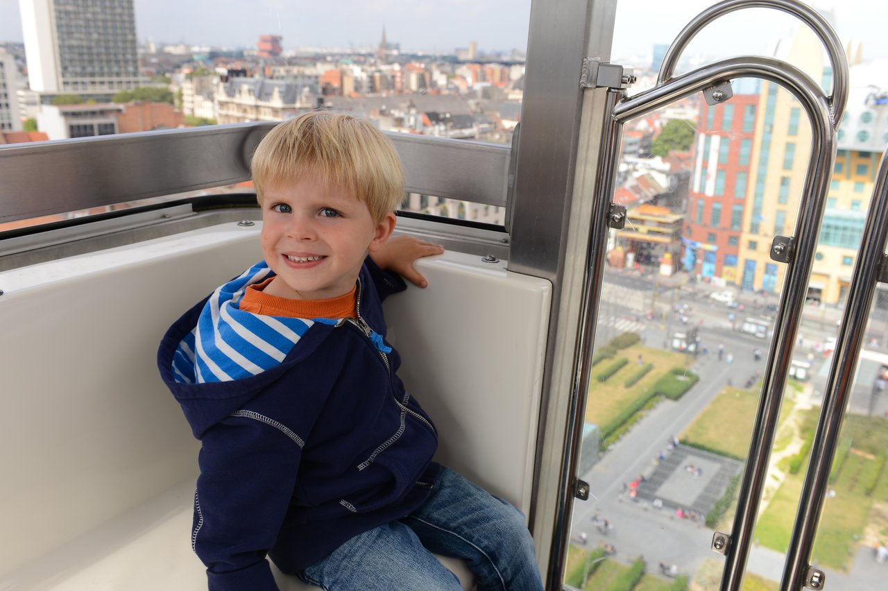 A young child sits inside a Ferris wheel cabin, smiling with a city view in the background.
