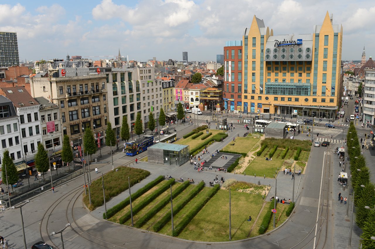 A city square with green spaces, tram tracks, and people walking.