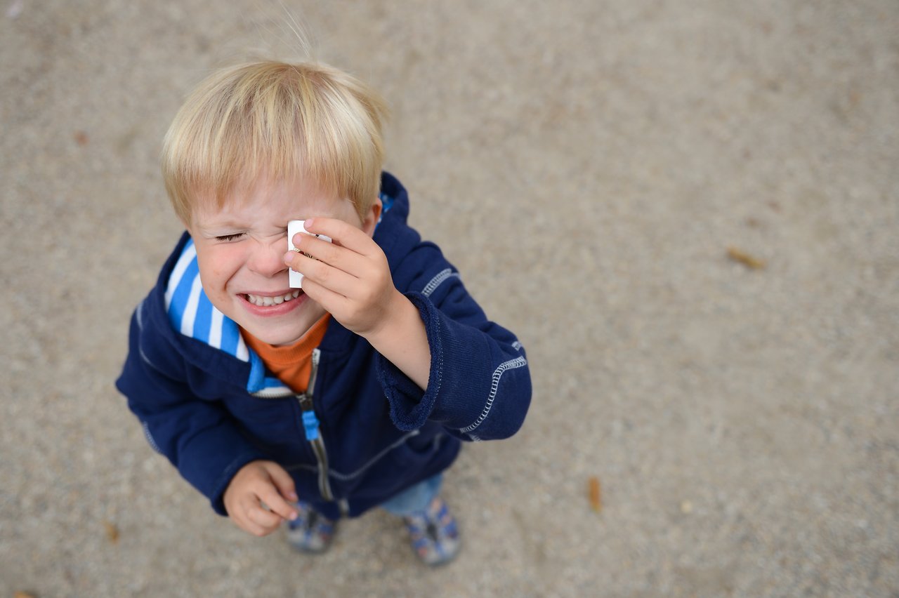 A young child in a blue jacket squints and wipes their eye with a tissue while standing outside.