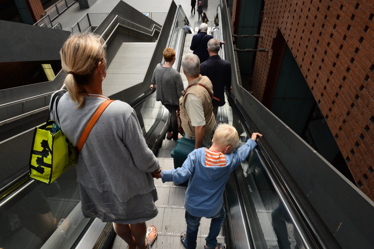 A woman and a child hold hands while descending an escalator with other people.