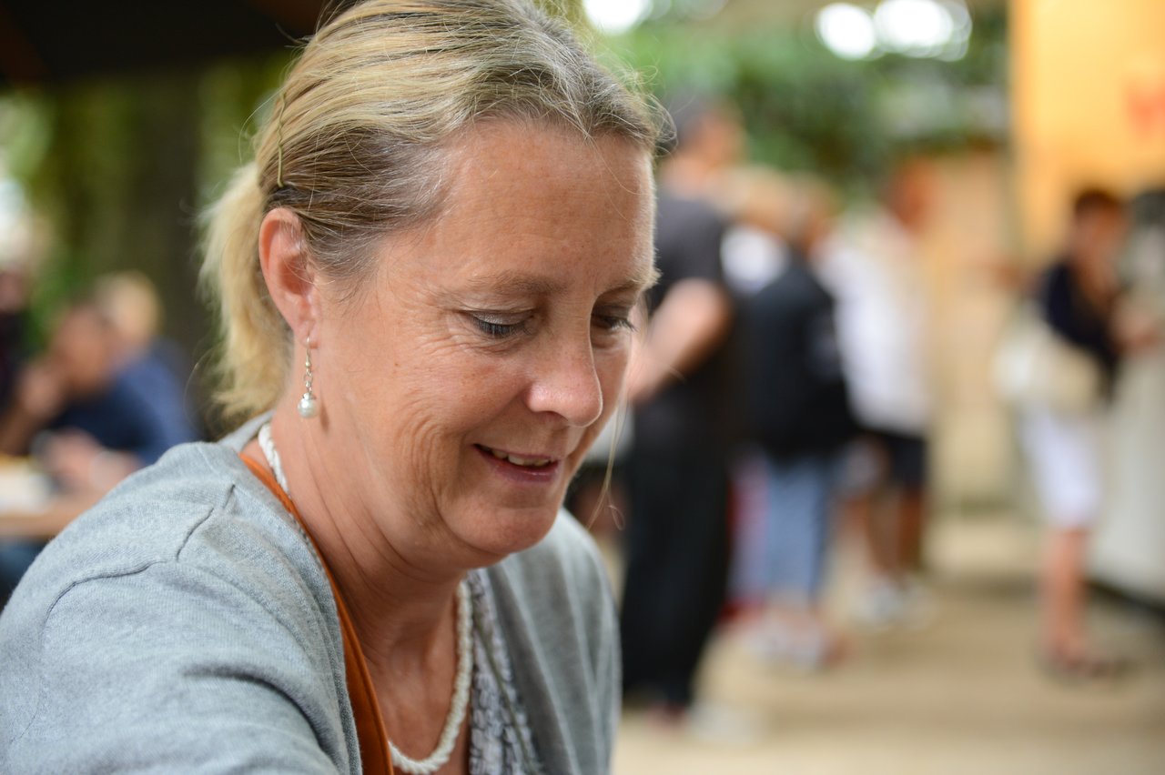 A smiling woman with blonde hair looks down, wearing a gray sweater and pearl earrings in a busy outdoor setting.