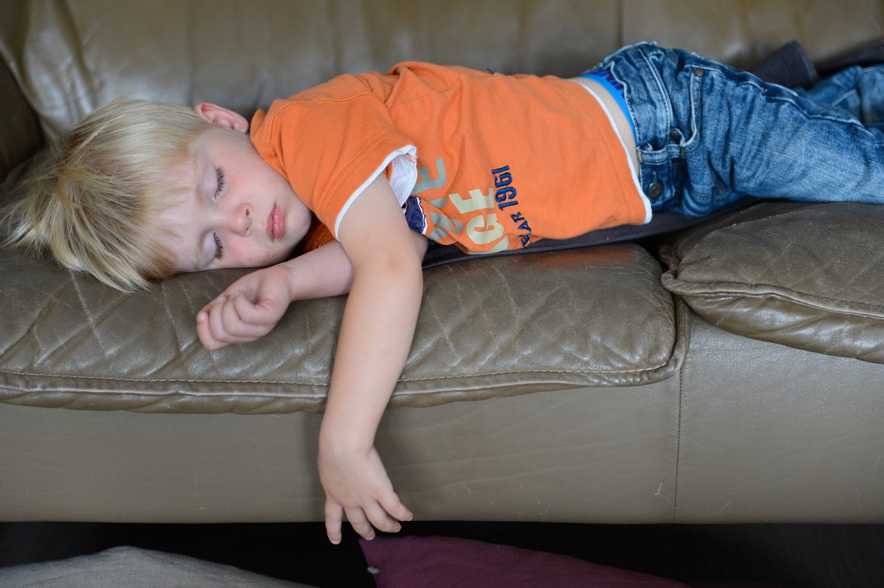 A young child in an orange shirt and jeans sleeps on a brown leather couch, resting with one arm hanging down.