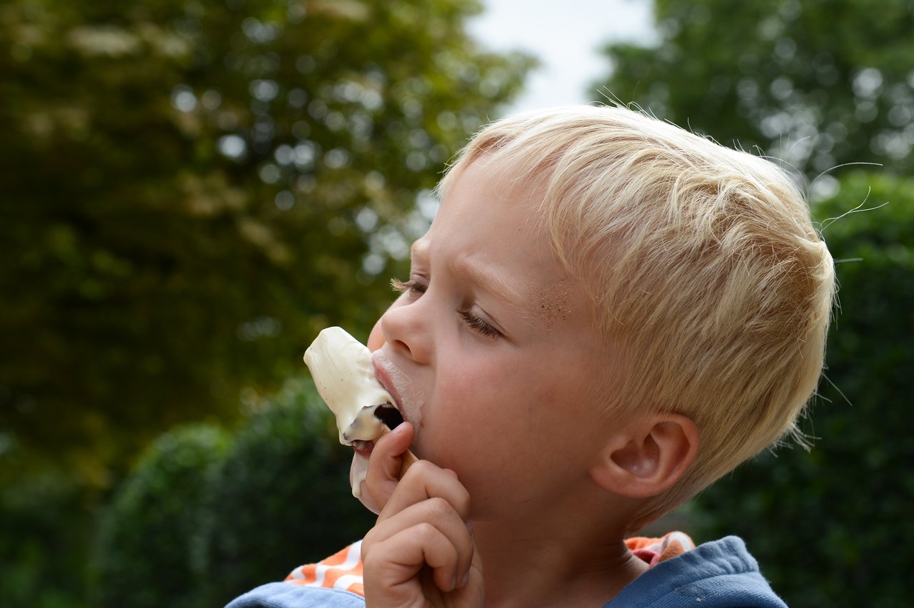 A young child with blonde hair eats a melting ice cream bar, with some ice cream on their face.