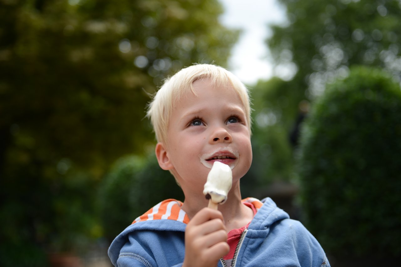 A young child in a blue hoodie holds an ice cream bar and looks up with a messy mouth.