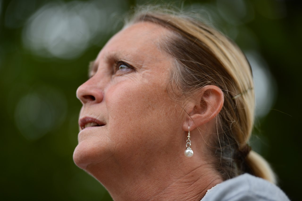A woman with blonde hair and pearl earrings looks up with a thoughtful expression against a blurred green background.