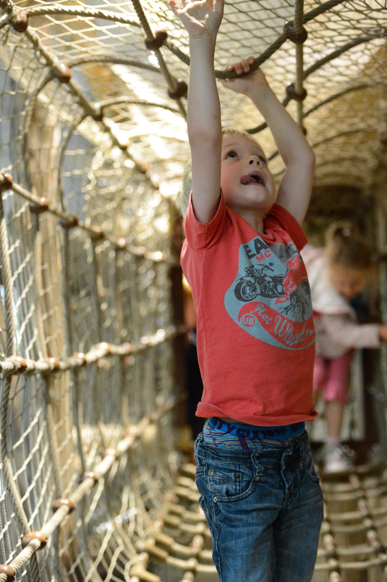 A child in a red shirt hangs from rope in a netted play structure, reaching upward with both hands.