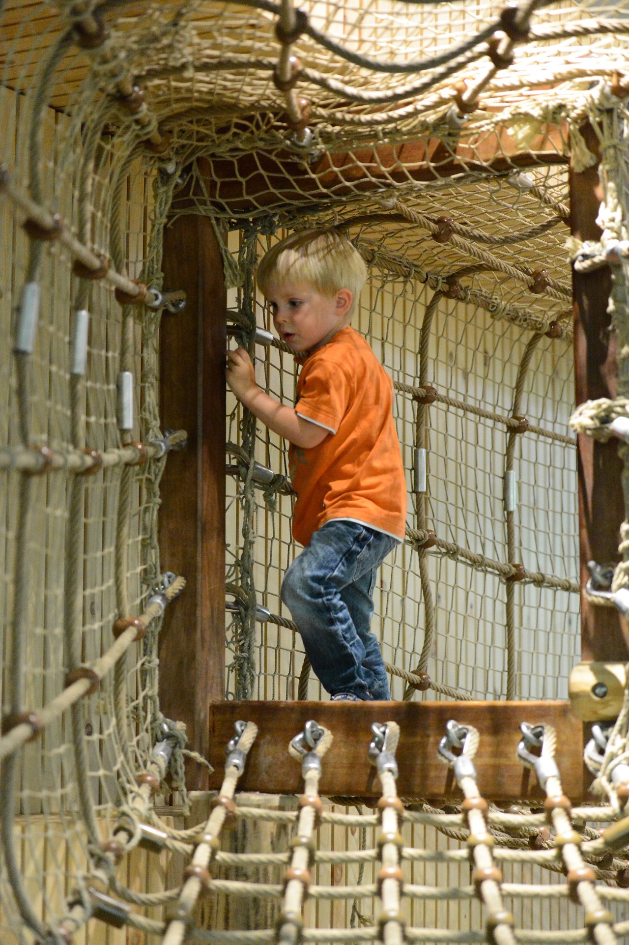 A young child in an orange shirt climbs through a rope tunnel at a zoo play area.
