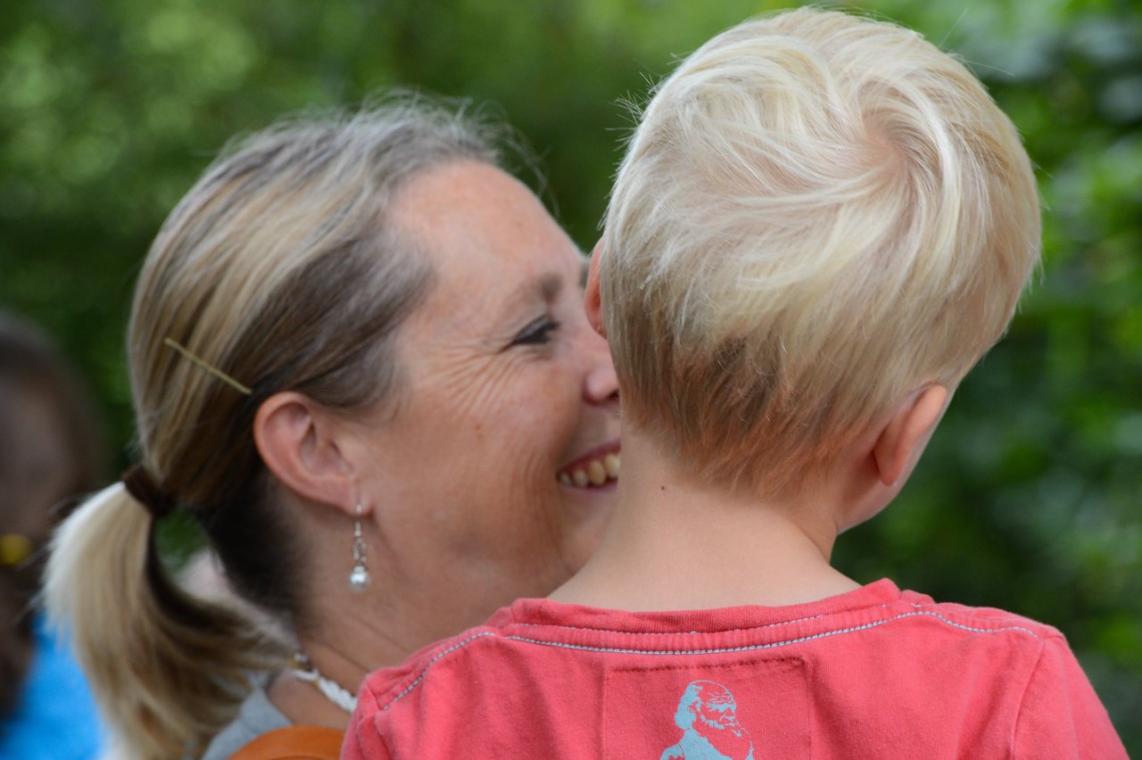 A woman smiles while holding a young child with blonde hair, both facing each other closely.