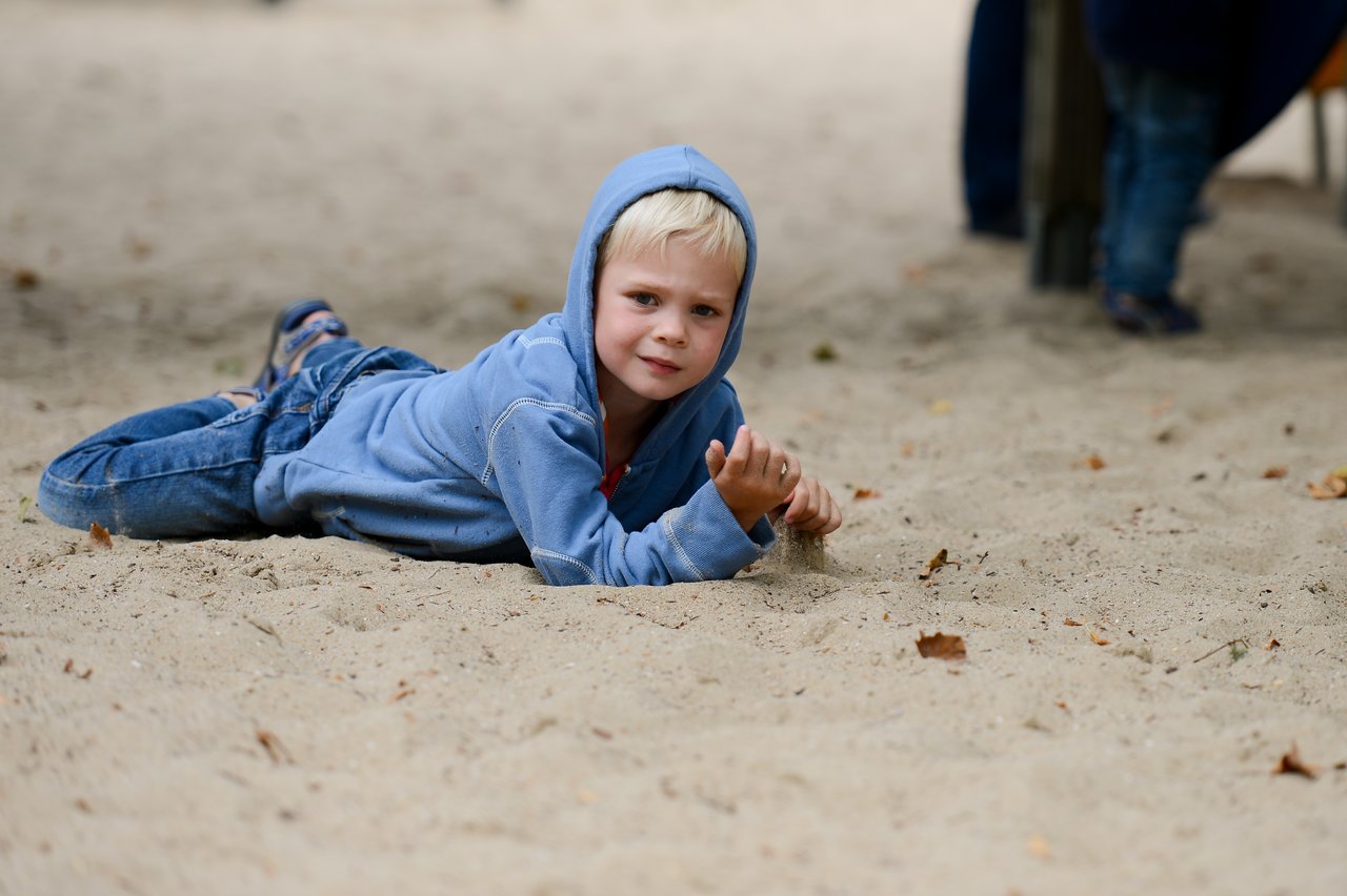 A child in a blue hoodie lies on the sand, propped on one arm, holding a small stick.