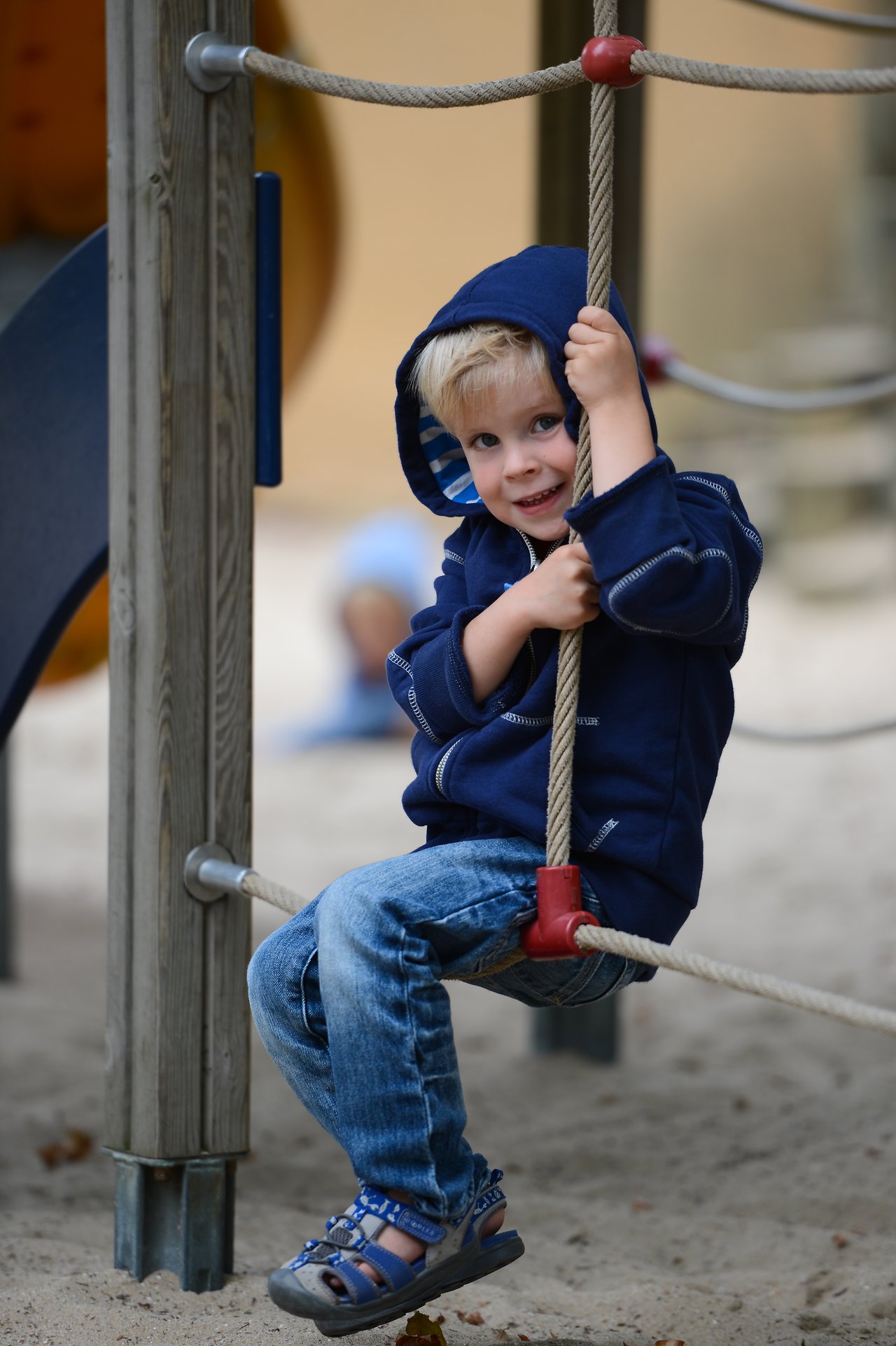 A young child in a blue hoodie sits on a rope in a playground, holding on and smiling.