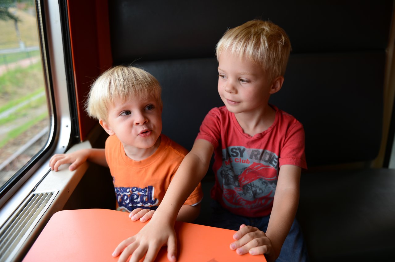 Two young children sit on a train, one talking excitedly while the other listens and reaches across the table.