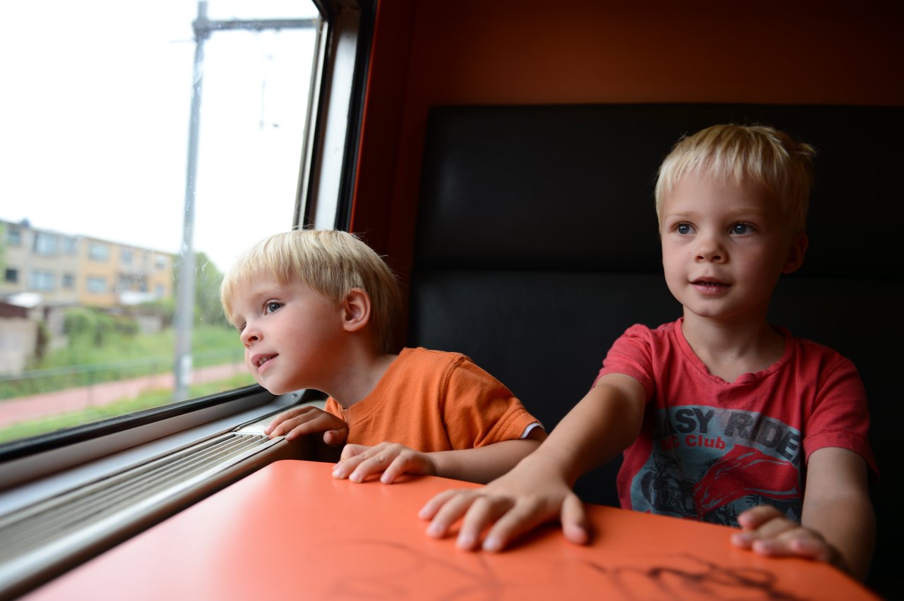 Two young children sit on a train, one looking out the window while the other rests a hand on the table.