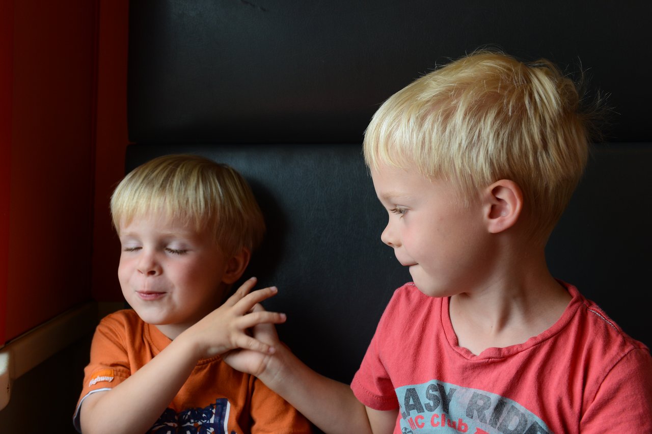 Two young children sit on a train, one playfully touching the other's hand while smiling.