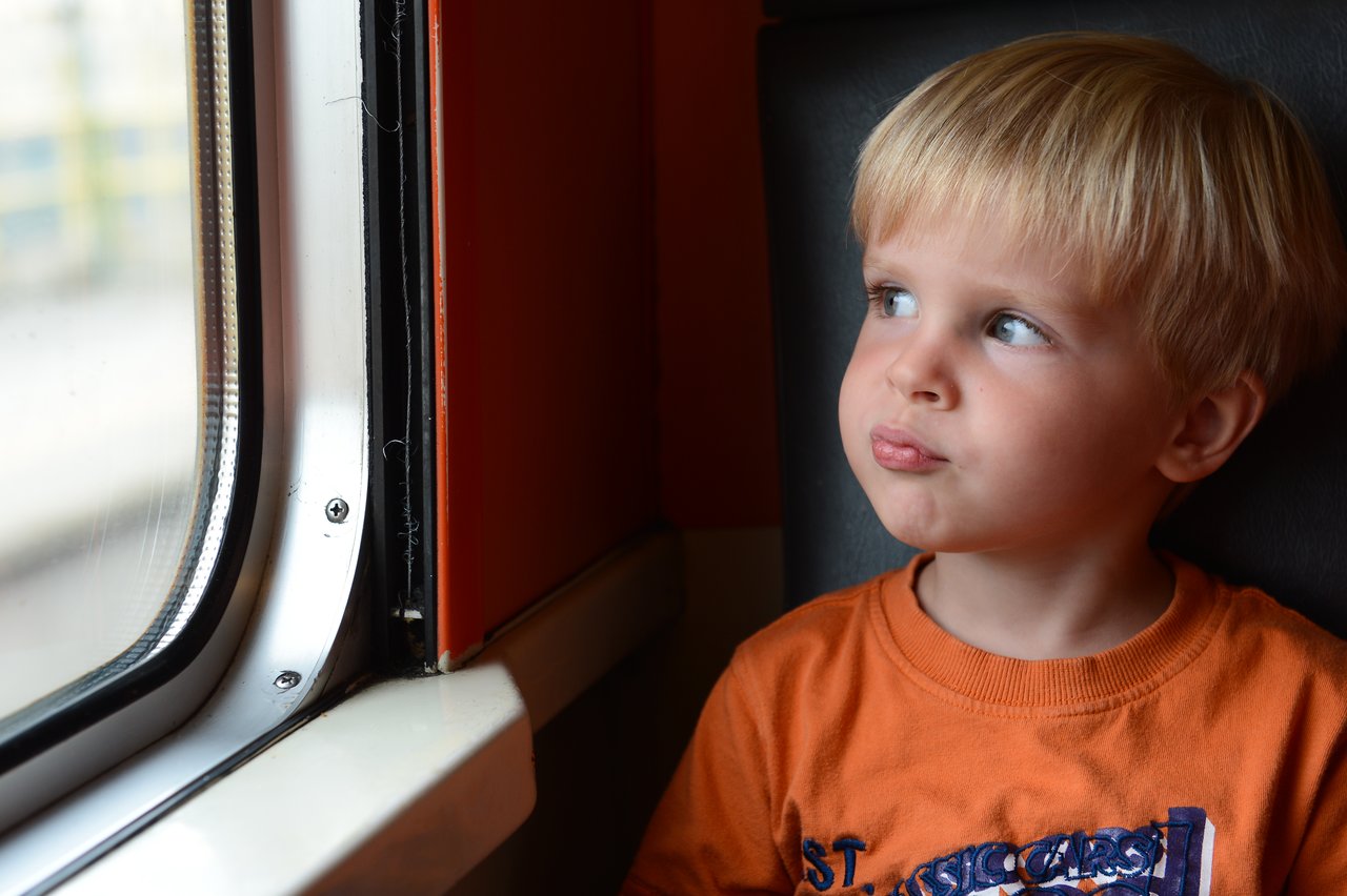 A young child in an orange shirt sits by a train window, looking outside with a thoughtful expression.