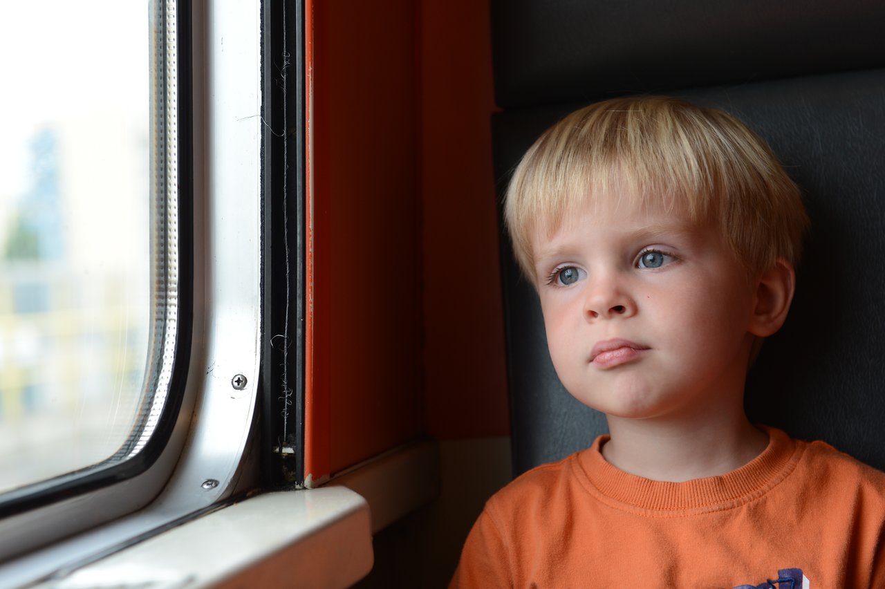 A young child in an orange shirt sits by a train window, looking outside with a thoughtful expression.