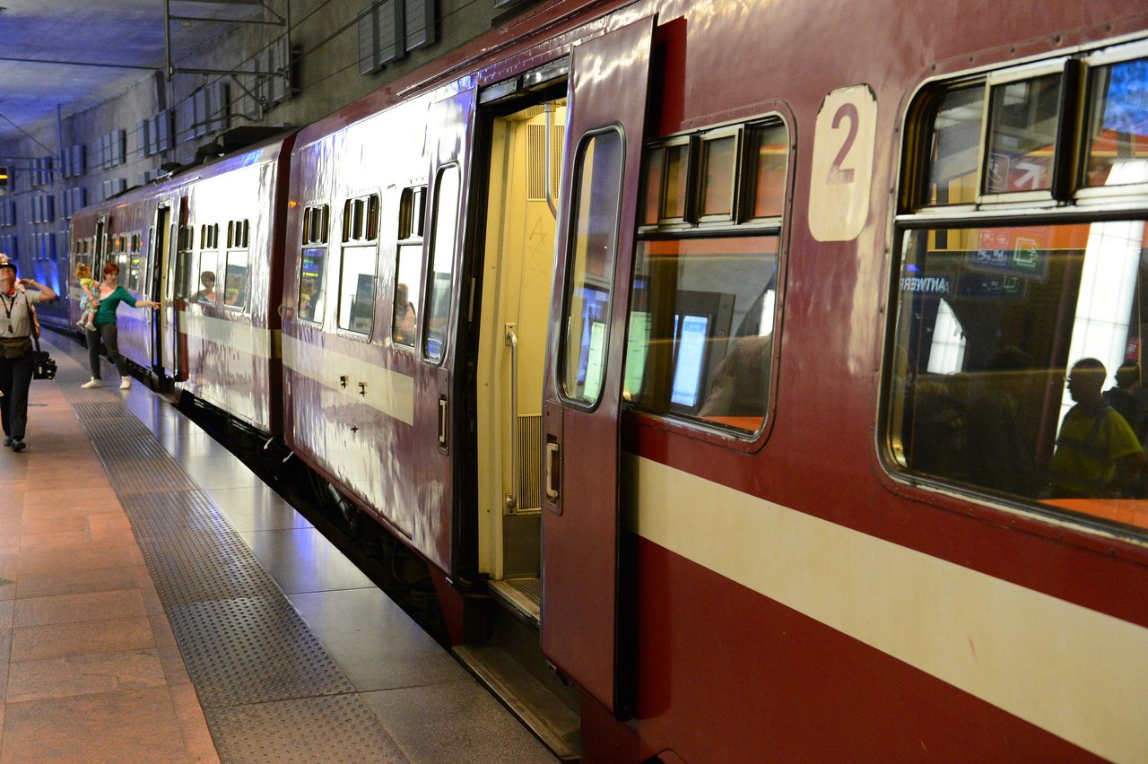 A red train is stopped at a station platform with passengers boarding and exiting through open doors.
