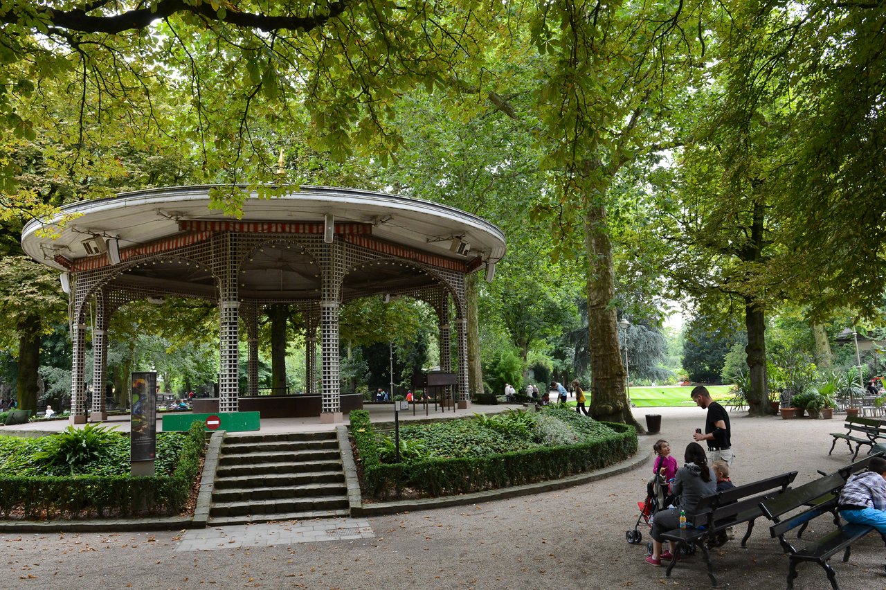 A gazebo in a green park with people sitting on benches and a man interacting with children nearby.