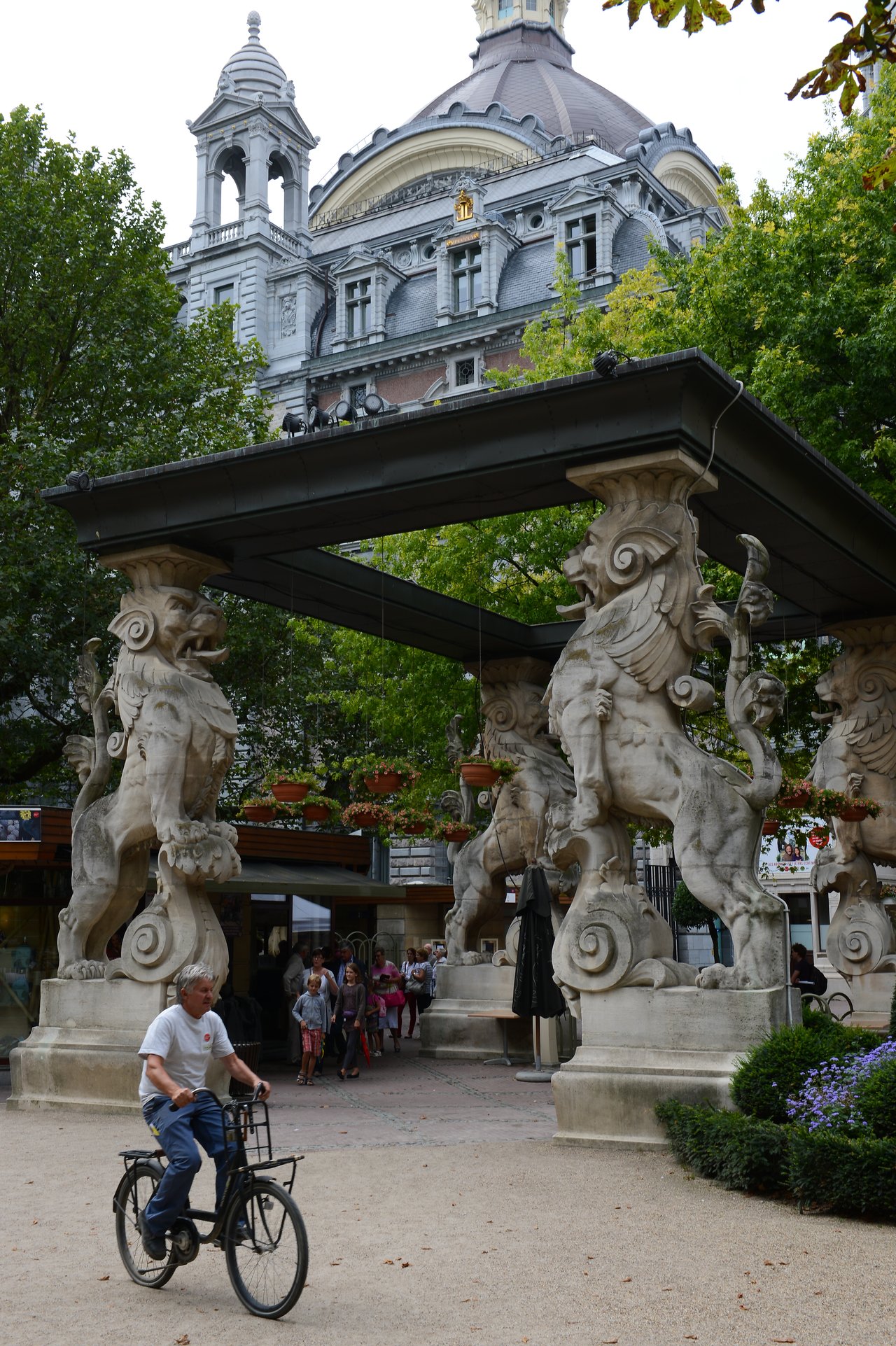 A man rides a bicycle past the entrance of Antwerp Zoo, featuring large stone animal sculptures.