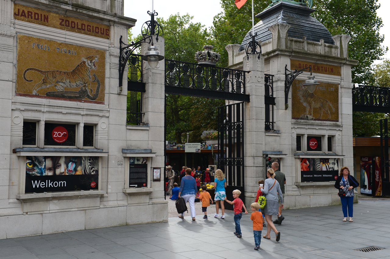 People, including children, walk through the entrance gate of Antwerp Zoo, which has decorative animal mosaics and signs.