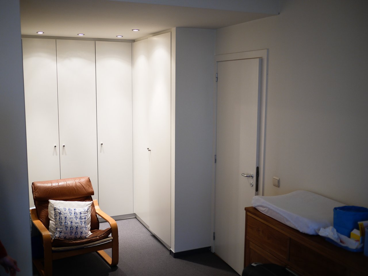 A small room with white wardrobes, a brown armchair with a pillow, and a wooden dresser with baby items.