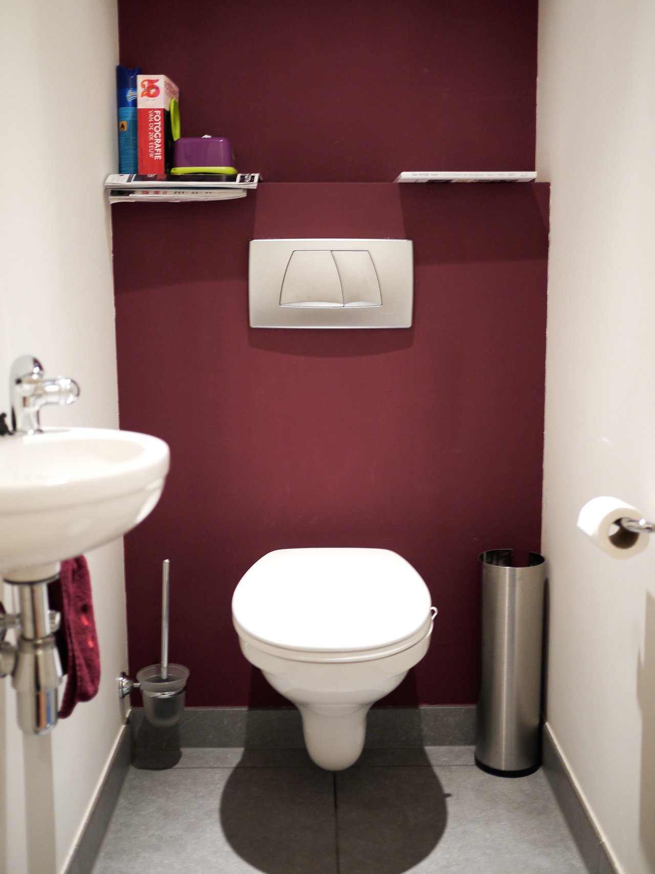 Small bathroom with a wall-mounted toilet, sink, and shelves holding toiletries against a dark red wall.