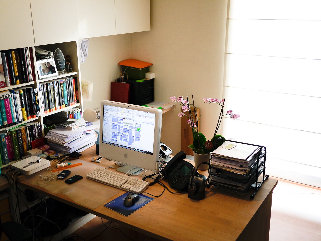 A home office with a computer, books, and office supplies on a wooden desk near a window with blinds.