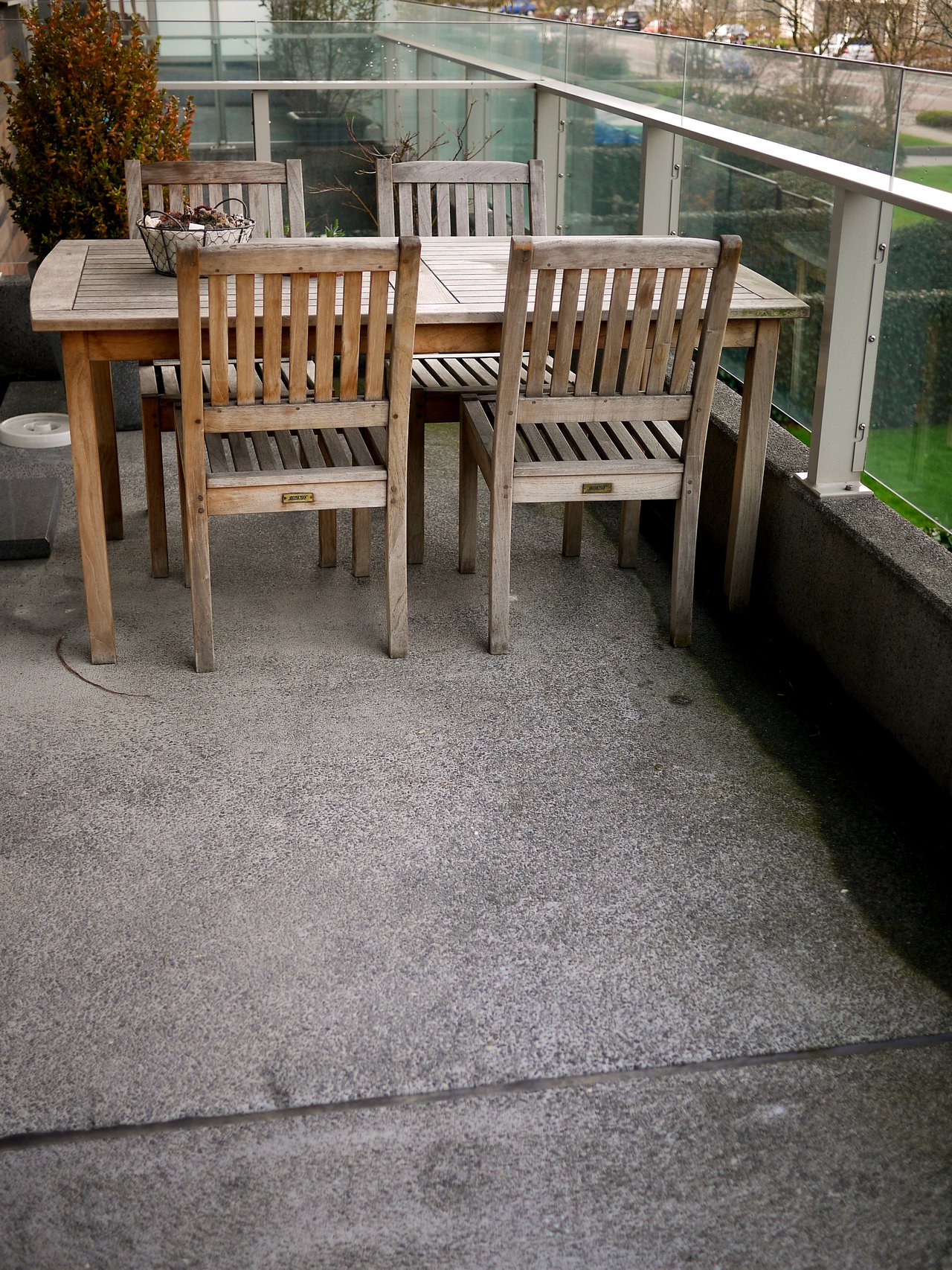 Wooden table with four matching chairs on a concrete balcony with glass railings, overlooking a street and greenery.