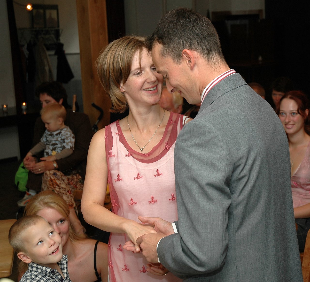 A smiling couple holds hands and looks at each other closely, surrounded by seated guests in a warmly lit room.