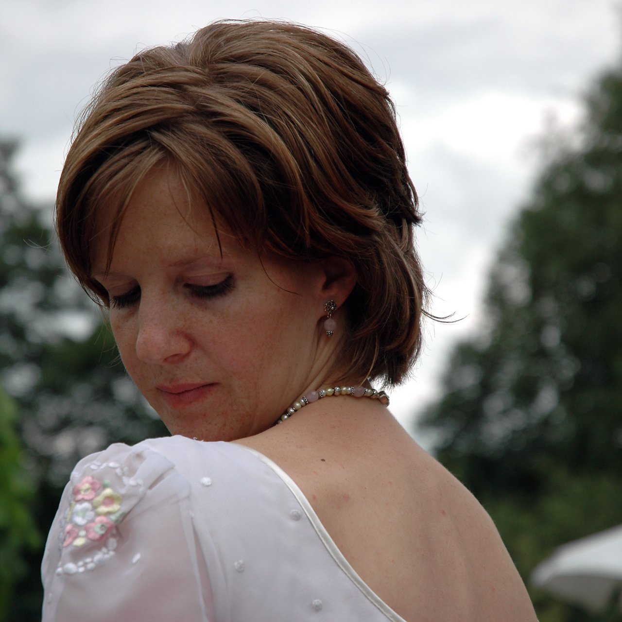 A woman in a white dress with floral details looks down, wearing a pearl necklace and earrings.