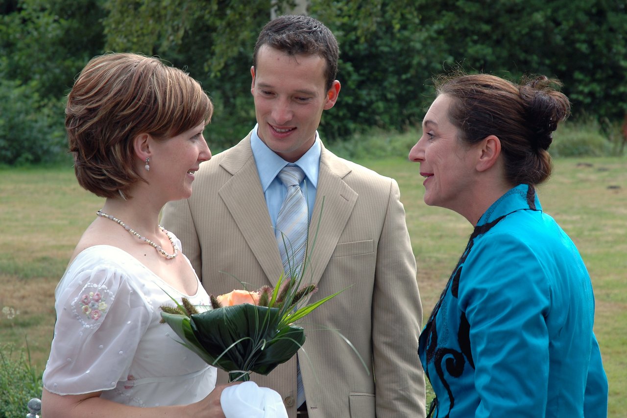 A bride holding flowers talks with a woman in a blue outfit while a man in a suit smiles.