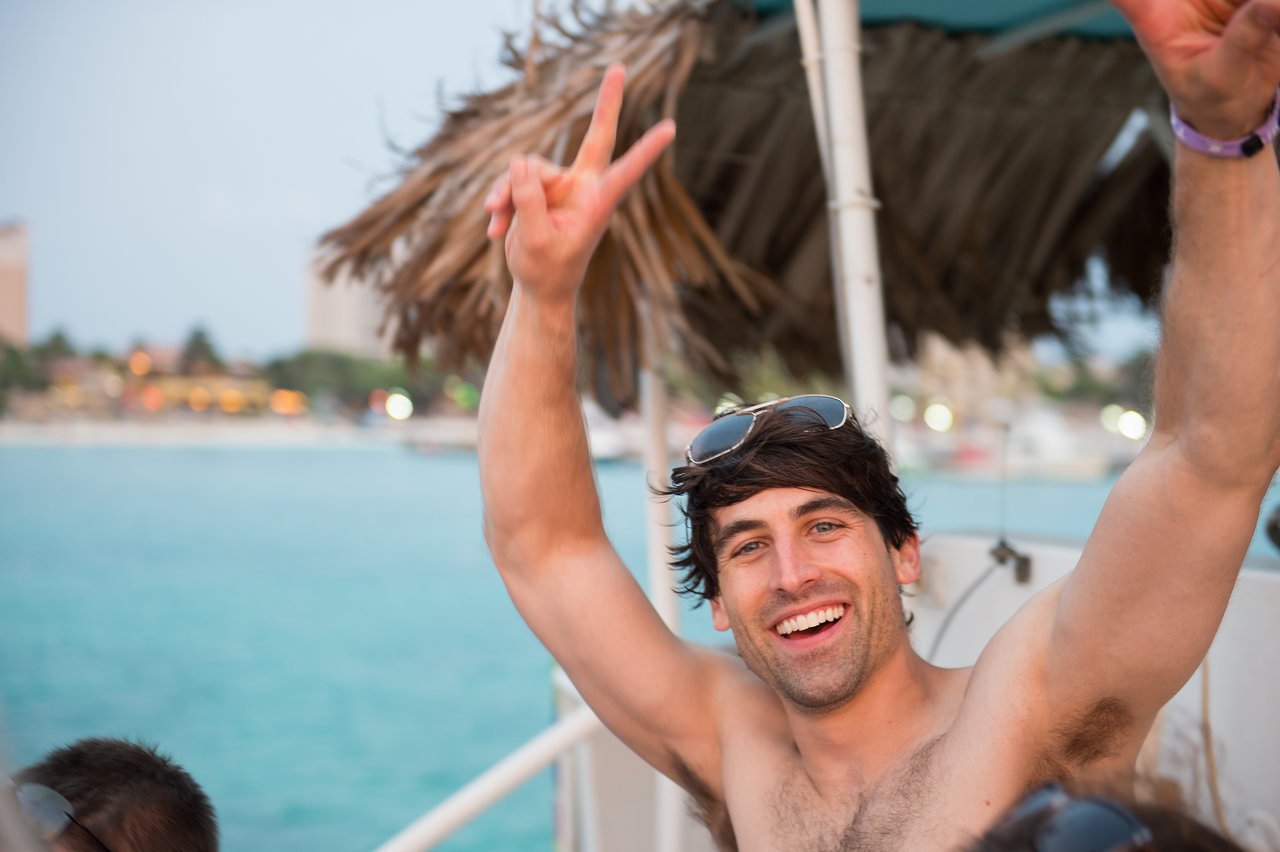 A smiling man on a boat raises his arms and makes a peace sign during a party.