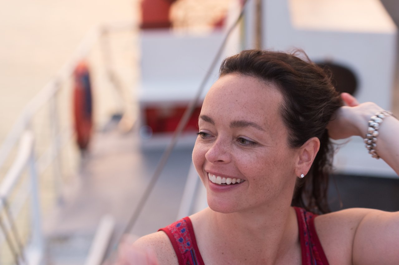 A smiling woman on a boat adjusts her hair, wearing a red top and a bracelet on her wrist.