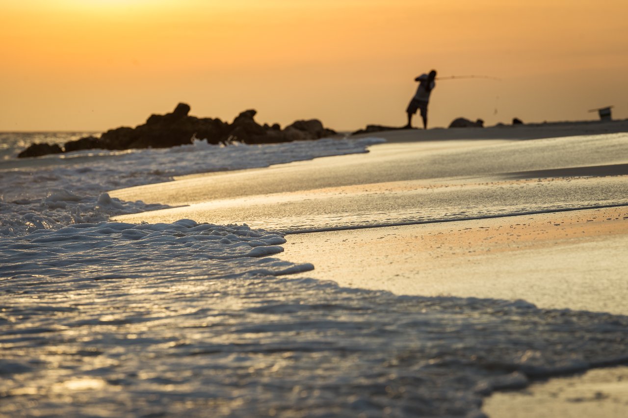 A person is fishing on the beach near the water's edge during sunset.