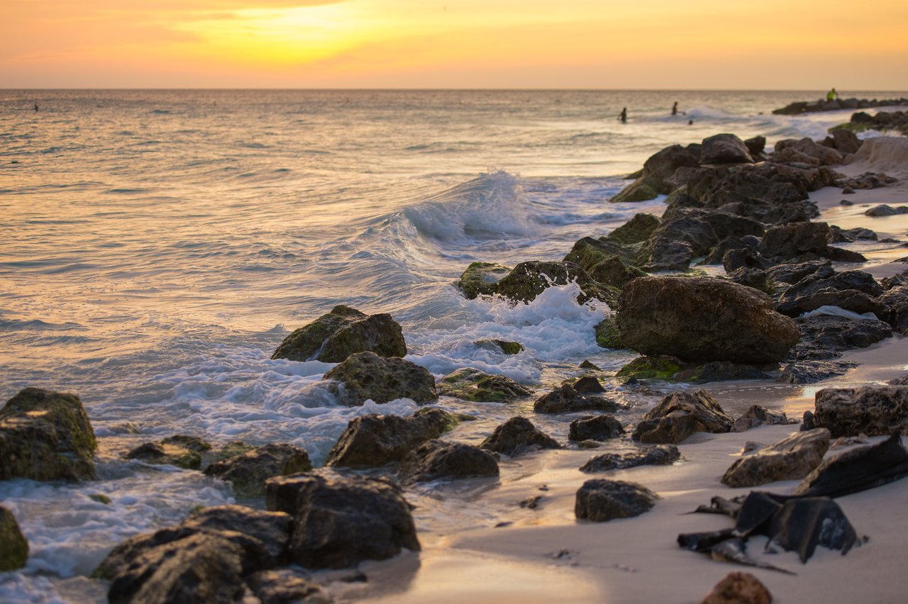 Waves crash against scattered rocks on a sandy beach at sunset, with a few people in the distance.