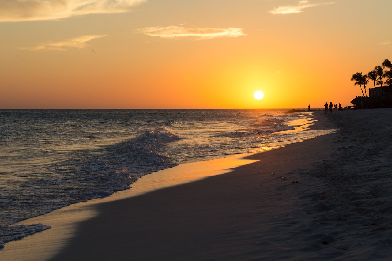 The sun sets over the ocean, casting light on the sandy beach with gentle waves and distant people walking.