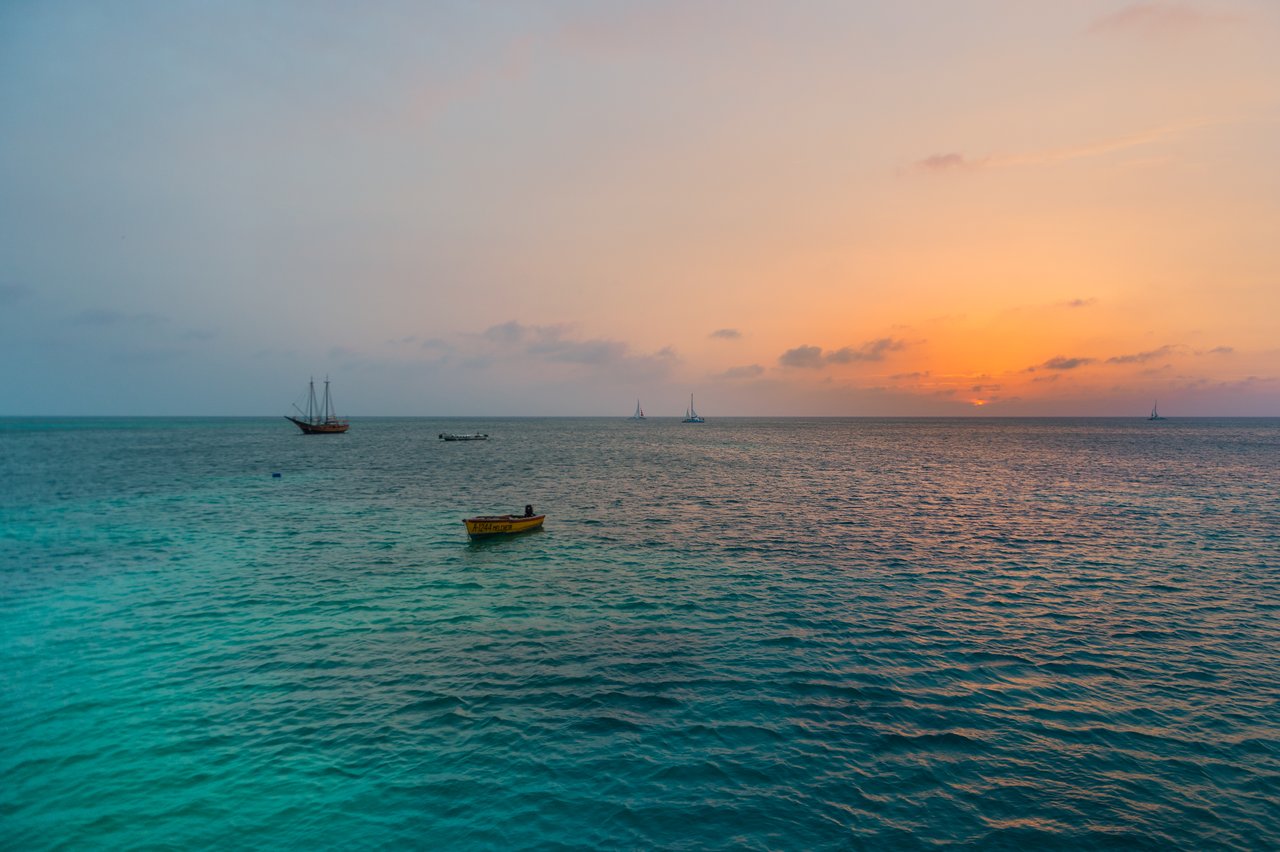 The sun sets over the ocean with several boats floating on the calm water.